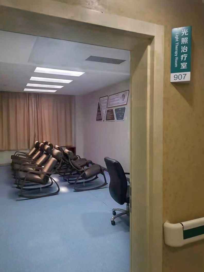 Doorway view into a hospital light-therapy room, with recliner chairs set beneath the skylight and a bilingual room sign confirming its use for daily light-therapy sessions.