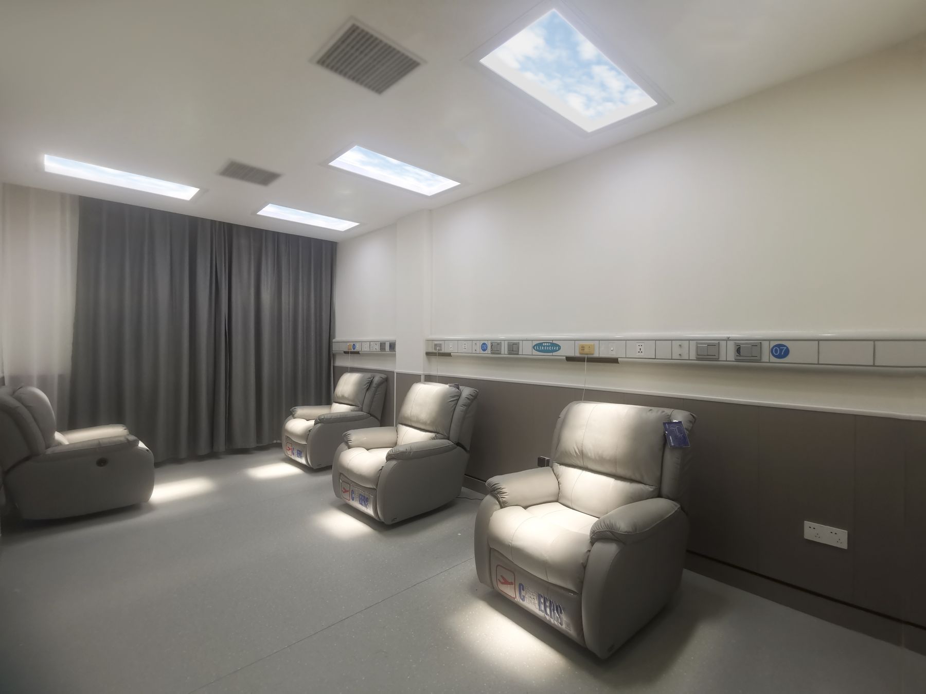 Static cloud skylight above a hospital light-therapy recliner room with curtained bays, giving patients access to a daily bright-light dose without leaving the ward.