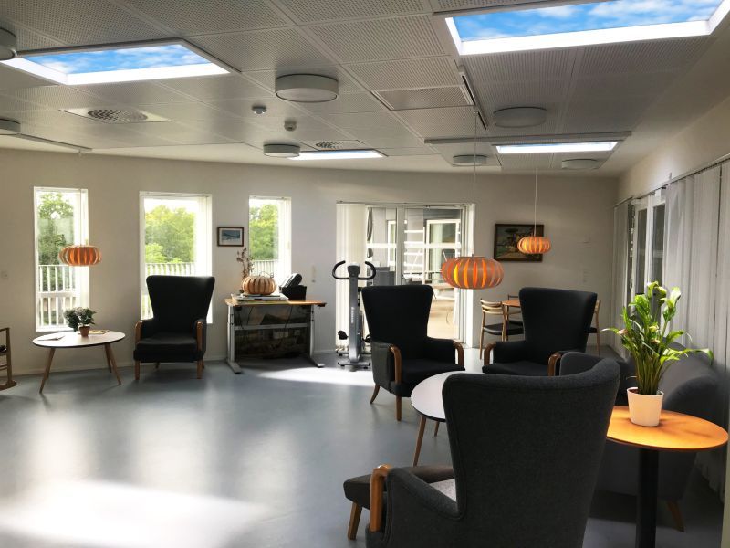Two static cloud skylights above a nursing home lounge furnished with wingback armchairs, giving residents a comfortable, naturally lit space to socialise and helping support a healthy day/night rhythm.
