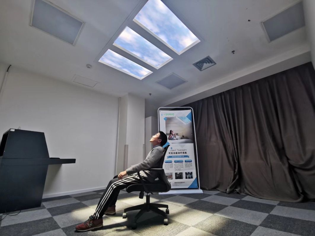 A patient reclines under the static cloud skylights during a light-therapy session in the same room, receiving a measured daily dose of bright light shown to ease seasonal depression and support circadian rhythm - the fixed, non-moving sky image keeps the light output steady and predictable for the length of the session.
