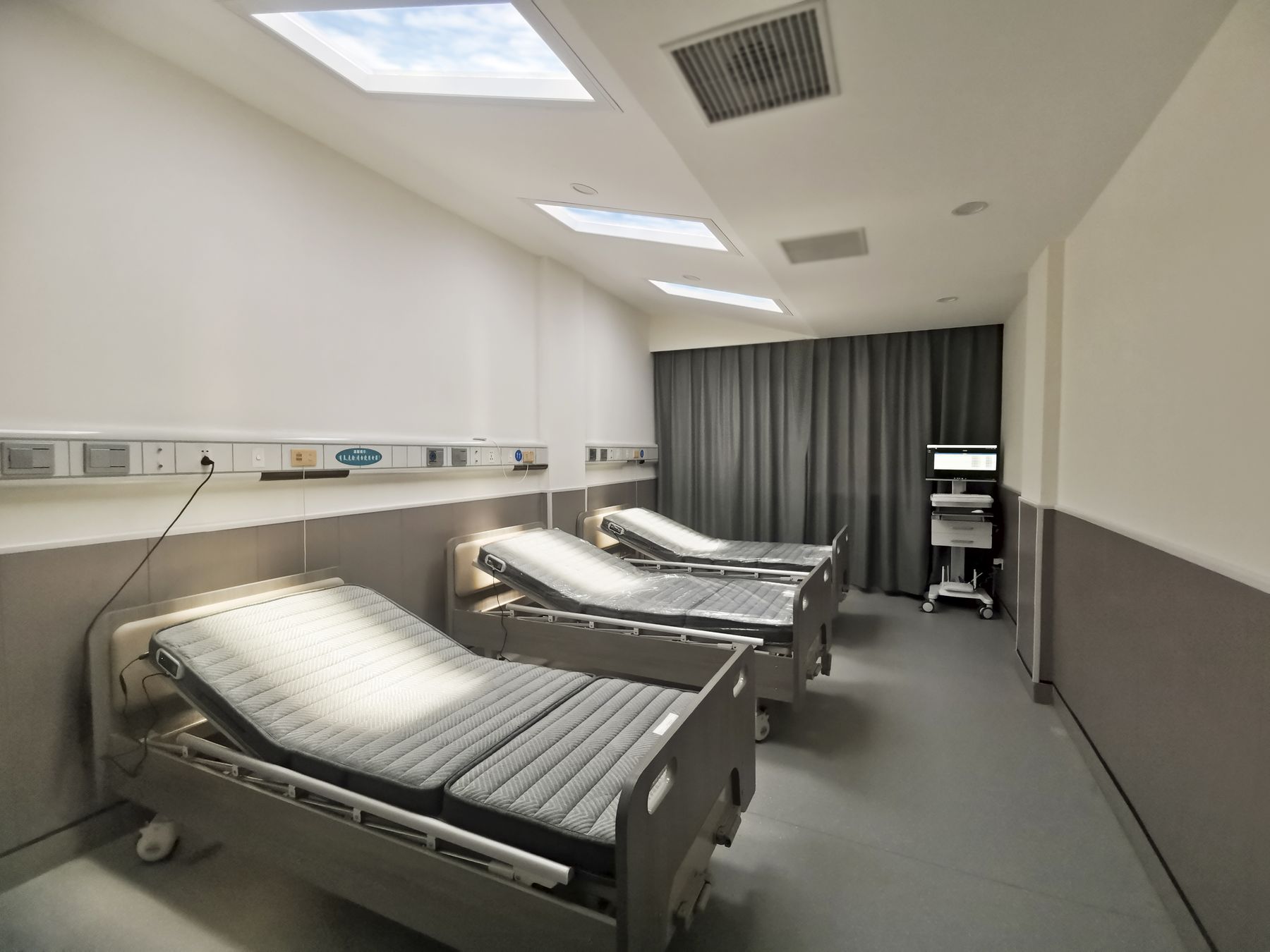 Two static cloud skylights above a two-bed hospital patient room, each bed angled toward the daylight feature to support rest and recovery.