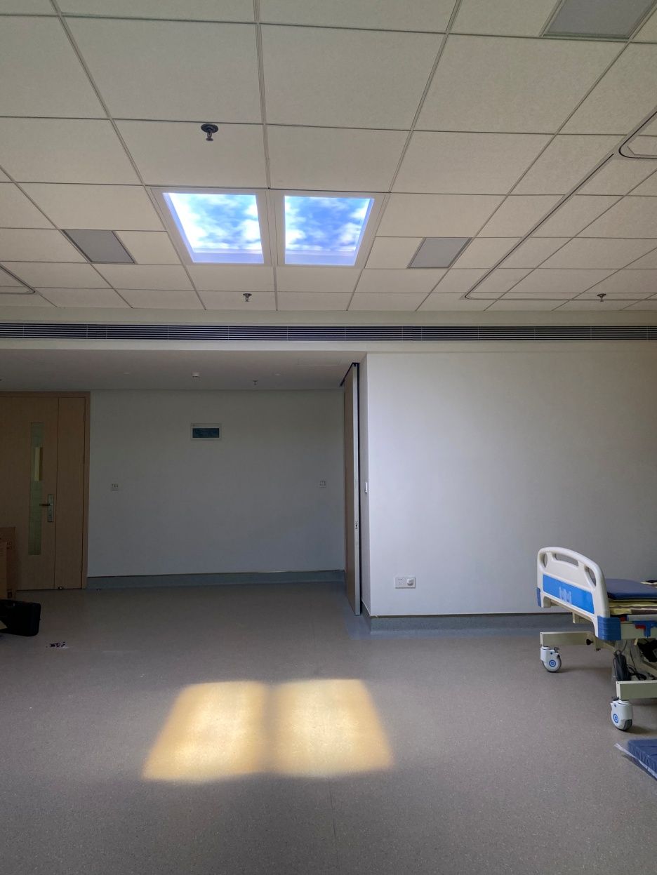 Second finished view of the rehabilitation ward, with dynamic sky skylights above the patient bay reinforcing the daylight theme throughout the department. As a dynamic sky panel, it also offers slow-moving clouds, an optional starry night mode after dark, and an adjustable colour temperature from crisp all-white daylight through to a warmer all-warm tone, letting the space be tuned through the day and evening rather than staying fixed like a static image.