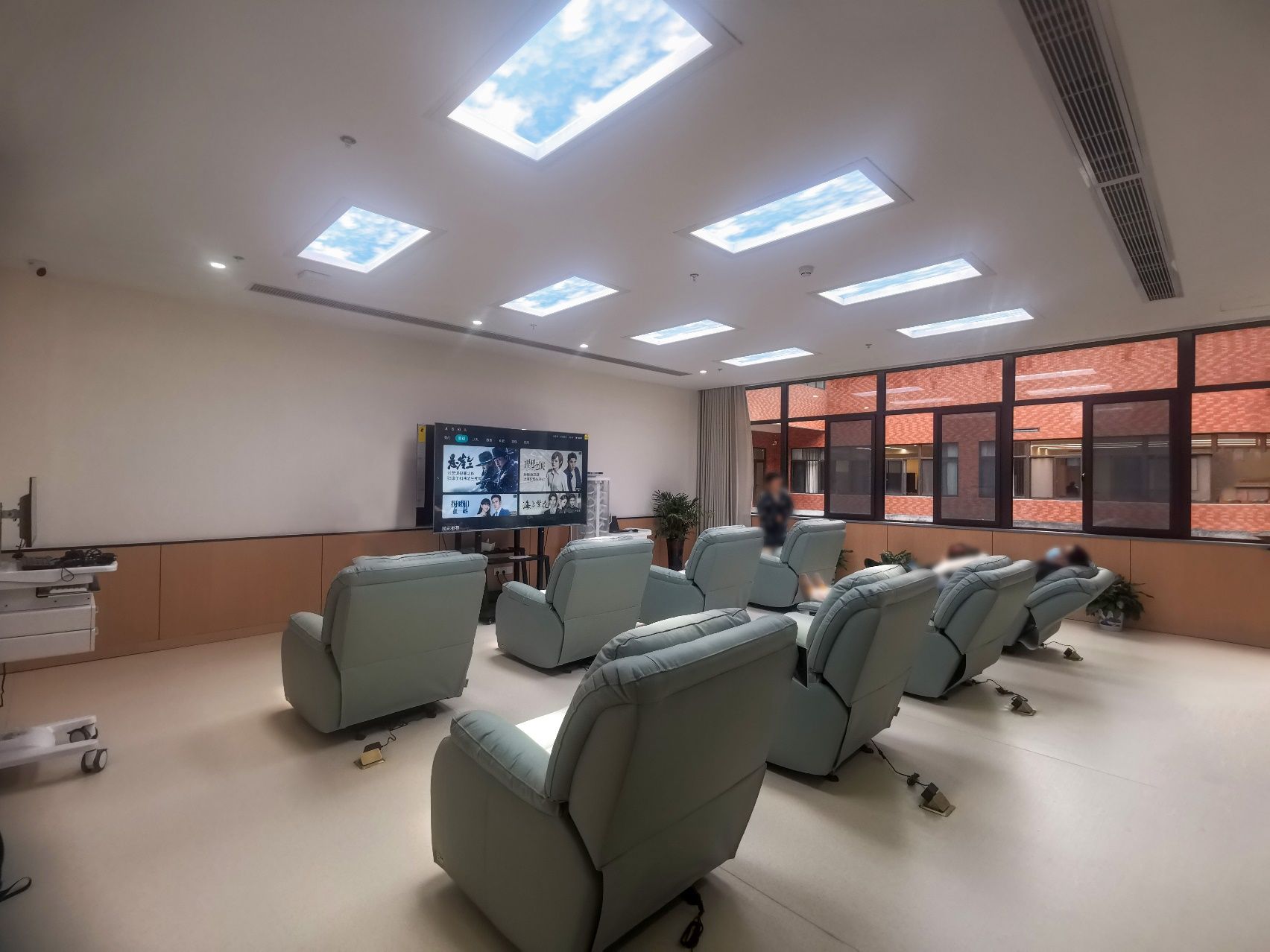 Six static cloud skylights above a hospital light-therapy theatre, giving a full row of patients a shared daily light-therapy session while facing a wall-mounted screen.