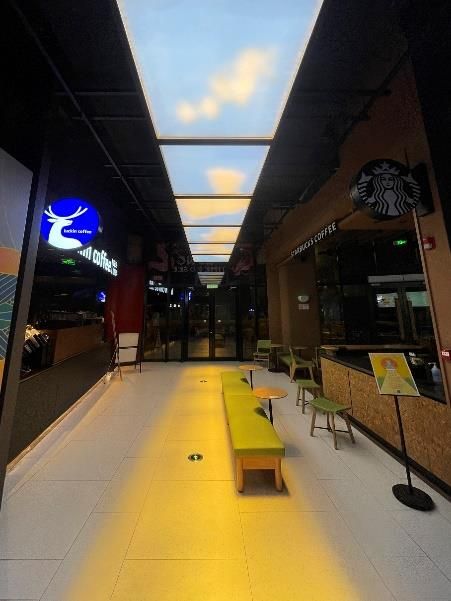 Grid of large scale 2400x1200 dynamic skylights installed down the centre of an indoor walkway within a shopping centre, bringing daylight down a windowless retail corridor past storefronts. As a dynamic sky panel, it also offers slow-moving clouds, an optional starry night mode after dark, and an adjustable colour temperature from crisp all-white daylight through to a warmer all-warm tone, letting the space be tuned through the day and evening rather than staying fixed like a static image.