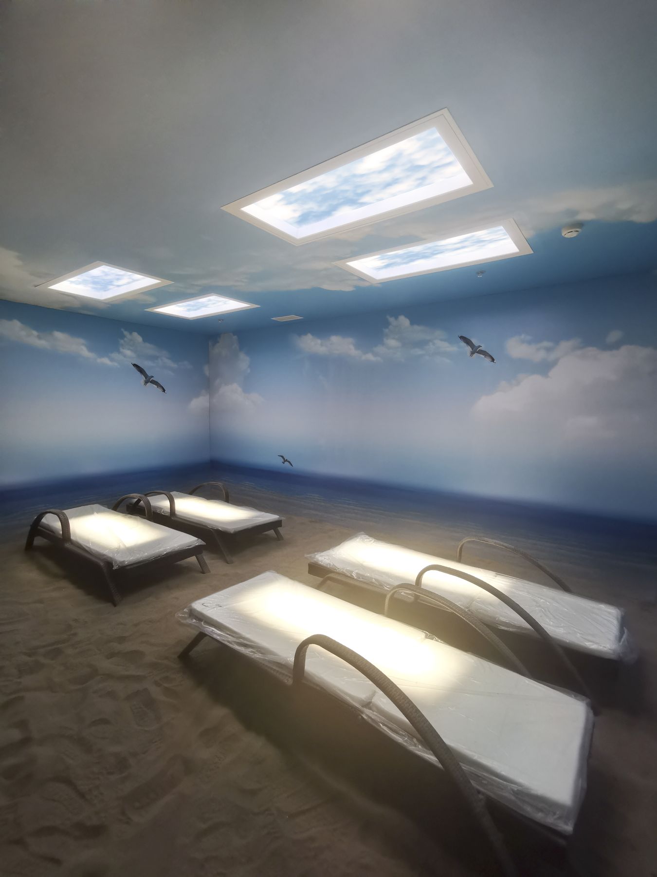 Three static cloud skylights above an immersive beach-themed light-therapy room, combining mural walls and real daylight above to support relaxation and stress reduction during treatment.