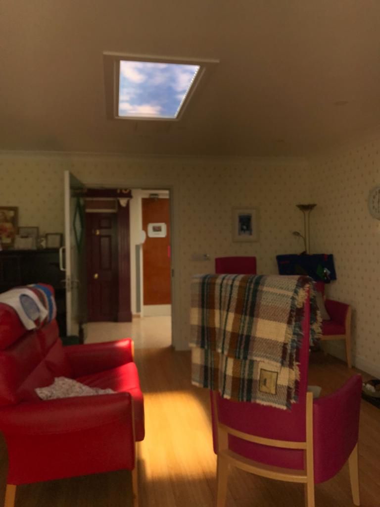 Evening view of the same dementia-care lounge, showing the skylight glowing above the seating area as part of the space's daily light routine. As a dynamic sky panel, it also offers slow-moving clouds, an optional starry night mode after dark, and an adjustable colour temperature from crisp all-white daylight through to a warmer all-warm tone, letting the space be tuned through the day and evening rather than staying fixed like a static image.